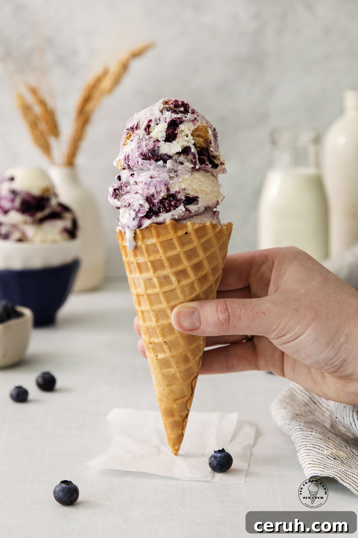 Blueberry Cheesecake Ice Cream Recipe — Creamy Homemade Treat 4 scoops of blueberry cheesecake ice cream in a waffle cone, held by a hand. Bottles of milk and cream are in the background