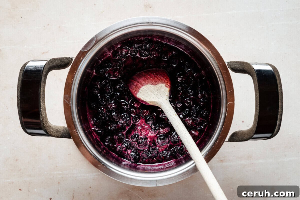 A pot filled with dark purple blueberry mixture being stirred with a wooden spoon, viewed from above on a light-colored surface.