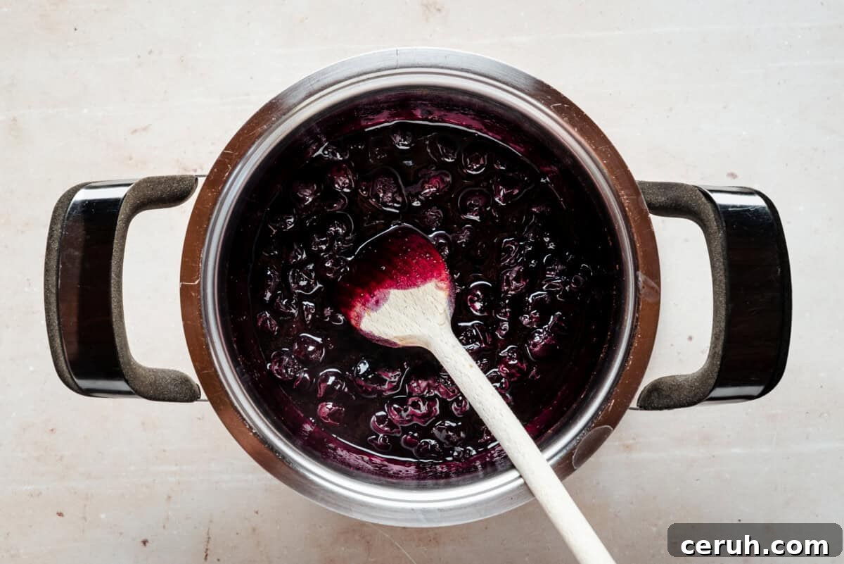 A pot of homemade blueberry compote being stirred with a wooden spoon, showing dark purple, glossy berries and syrup against a light background.