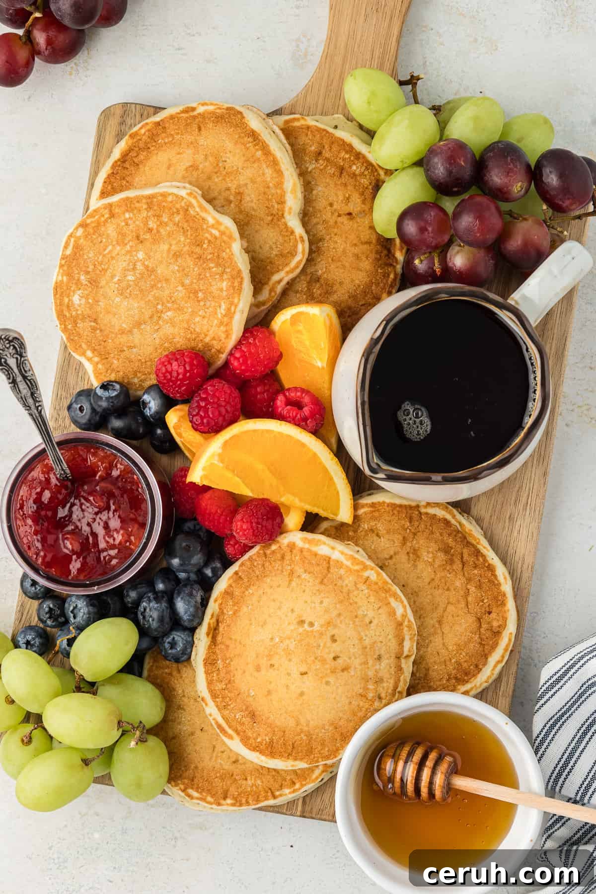 a wooden cutting board covered in buttermilk pancakes, a bowl of serving, a small bowl of jam with a spoon in it, a small bowl of honey, and fresh orange slices, raspberries, blueberries and green and red grapes
