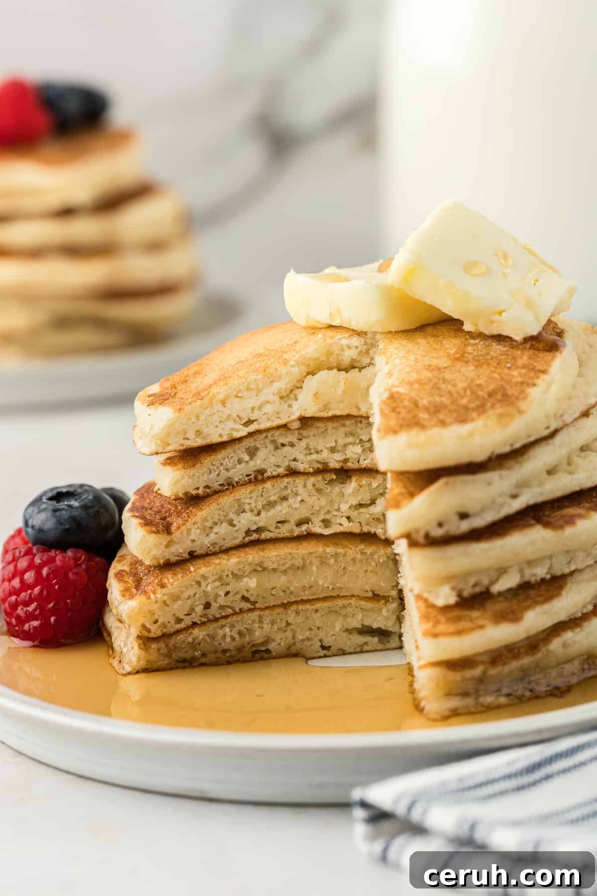 a white plate with 5 pancakes in a stack, with one large slice out of the entire stack, fresh blueberries and raspberries beside the pancakes, two slices of butter on top and syrup all over the plate