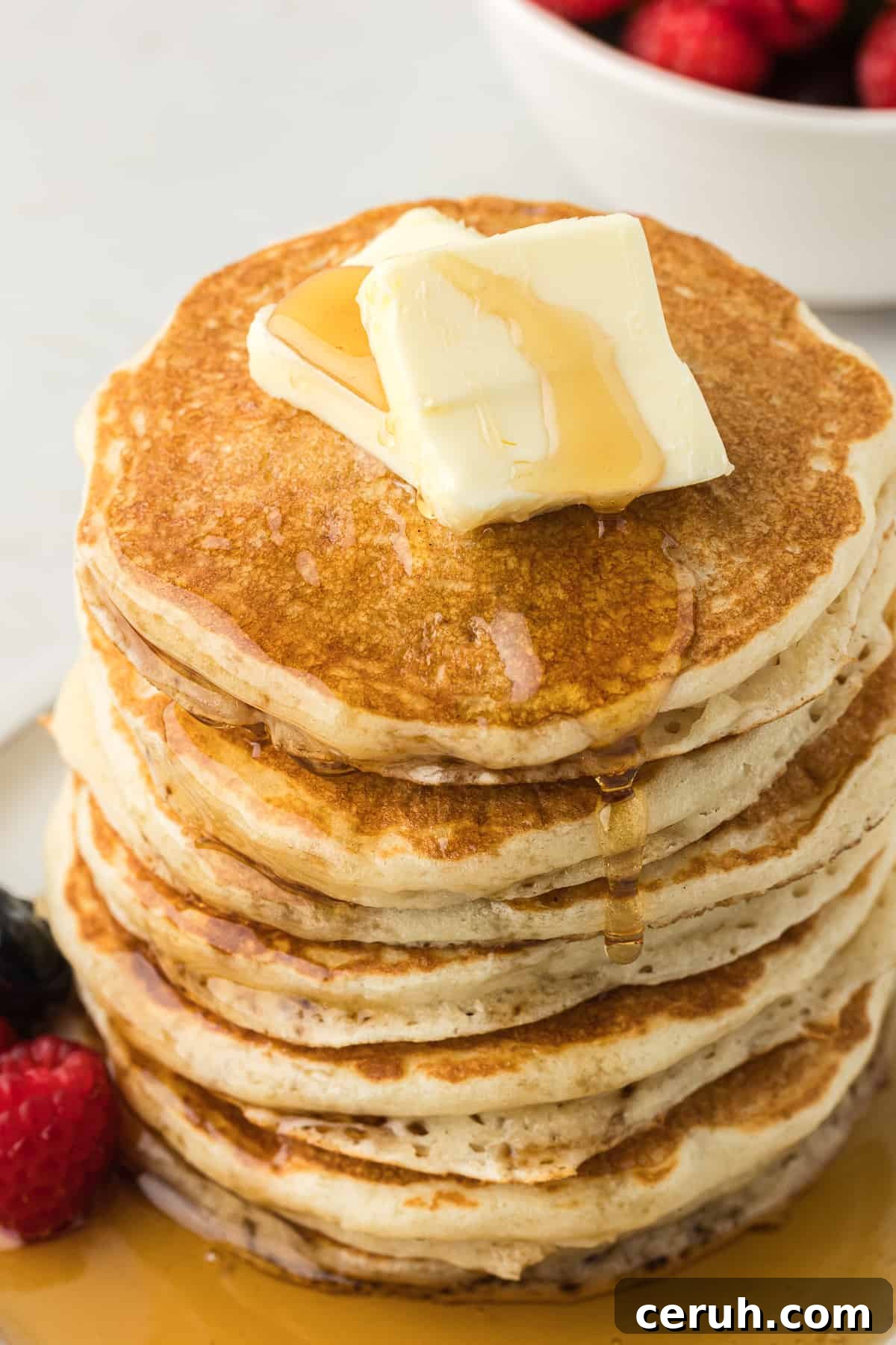 a tall stack of buttermilk pancakes on a white plate with two slices of butter on top and fresh blueberries and raspberries on the plate drizzled with maple syrup