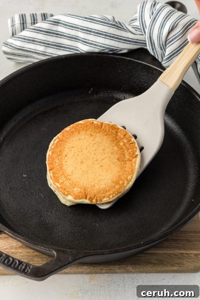 a buttermilk pancake being flipped with a spatula over a cast iron skillet that's held by the handle with a blue and white striped towel