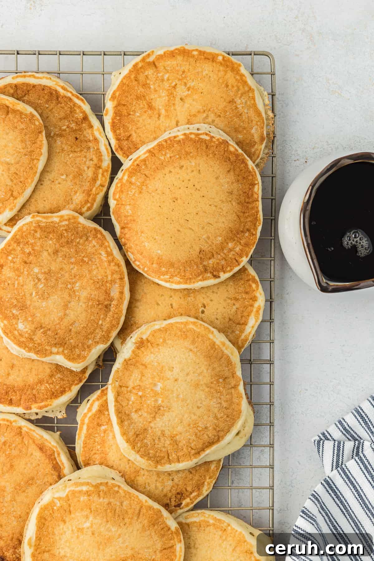 a wire rack covered in buttermilk pancakes with a bowl of syrup and blue and white striped towel beside it