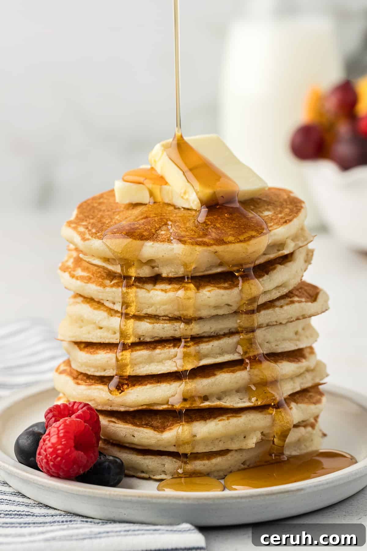 a tall stack of buttermilk pancakes on a white plate with two slices of butter on top and fresh blueberries and raspberries on the plate being drizzled with maple syrup