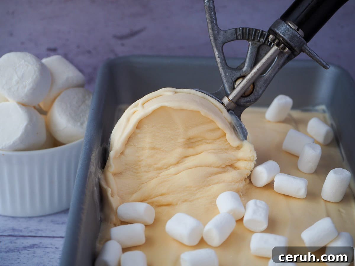 Scooping marshmallow ice cream, with mini marshmallows on top and bowl of marshmallows in the background.