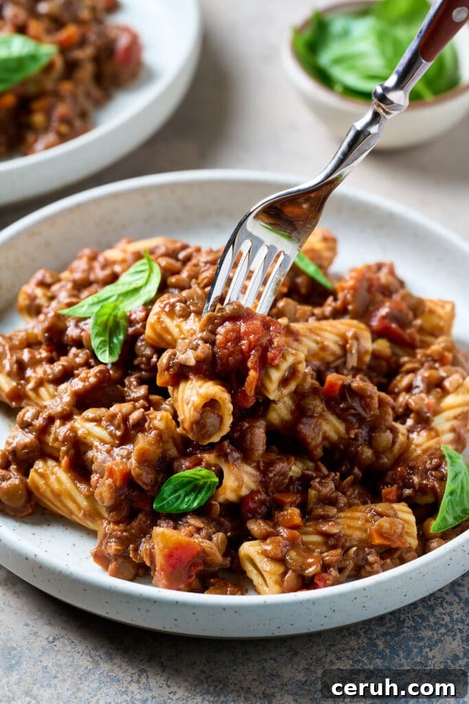 Fork lifting a bite of rigatoni pasta coated in lentil Bolognese from a plate.
