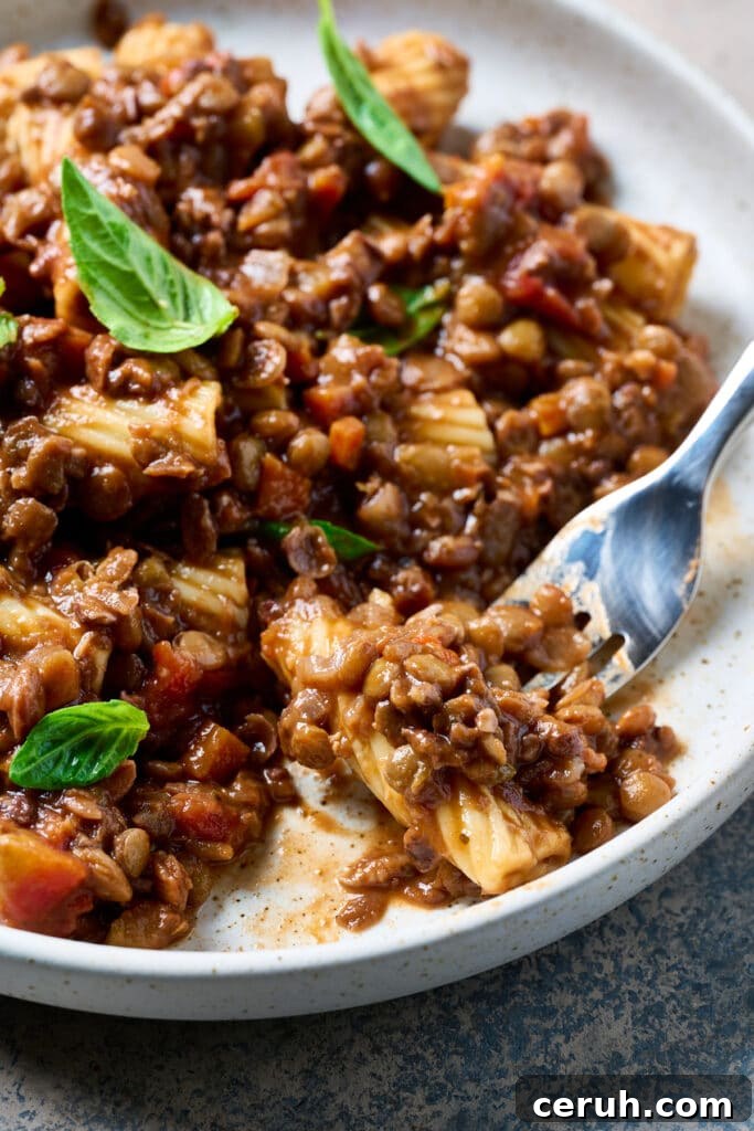 Close-up of rigatoni pasta with lentil Bolognese sauce, garnished with basil leaves, and a fork holding a bite.