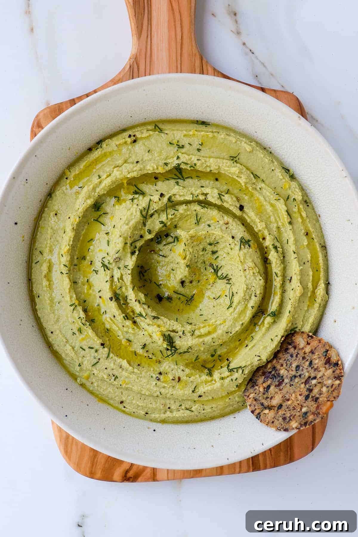 Lemon dill hummus in a bowl with cracker on wooden board.
