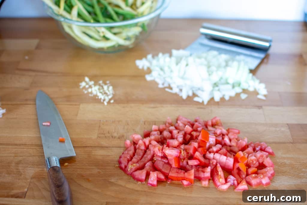 Chopped tomatoes, chopped onion, minced garlic, a chef's knife, and a scraper with a bowl of green beans and yellow wax beans.
