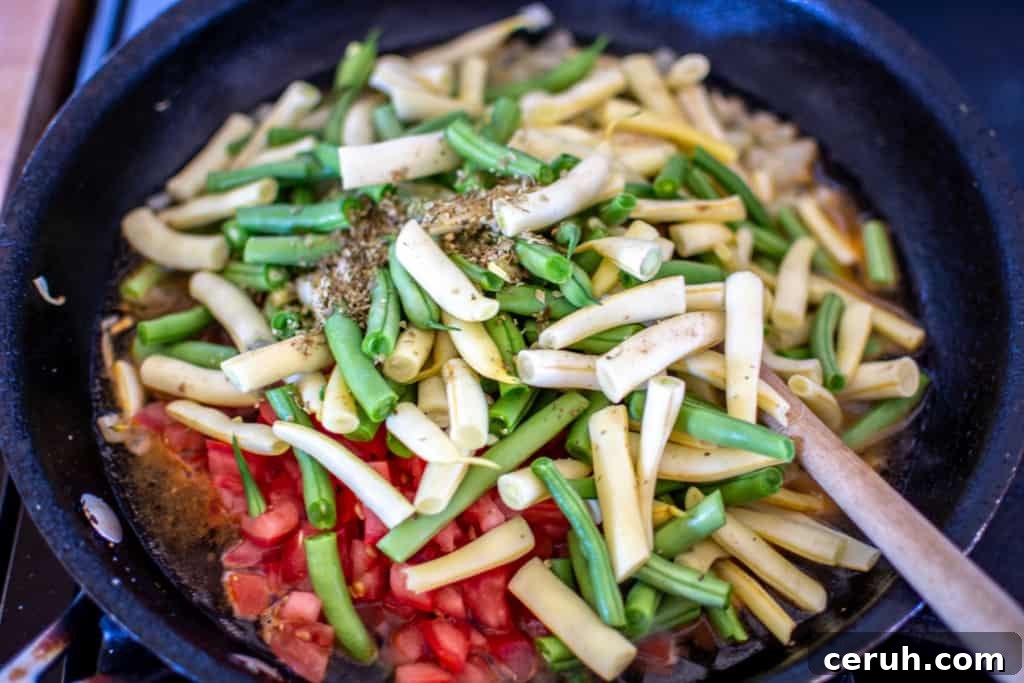 Chopped tomatoes, cut green beans and yellow wax beans, dried oregano, salt, and water in a black frying pan with a wood spoon.