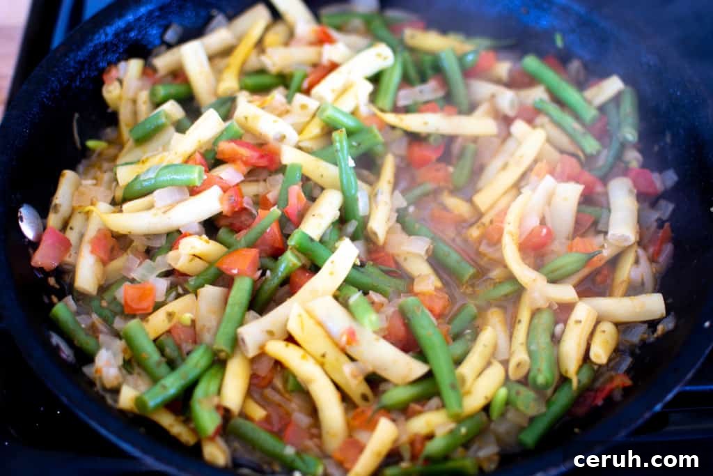A black frying pan with stewed green beans and yellow wax beans in it with chopped tomatoes all cooked with onions.