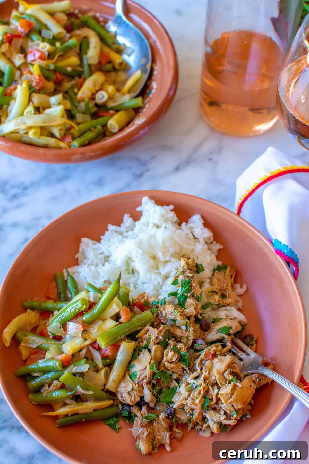 A brown plate filled with steamed rice, Chicken Picadillo, stewed green beans on a marble table next to a white napkin.