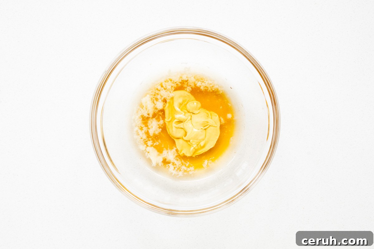 Vinegar, grated garlic, maple syrup and dijon mustard in a glass bowl viewed from above.