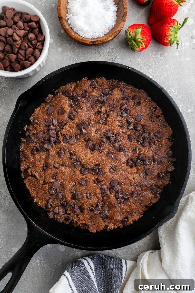 Gluten free chocolate chip cookie dough pressed into an 8 inch cast iron skillet with chocolate chips sprinkled on top surrounded by a small bowl of flaky sea salt and strawberries