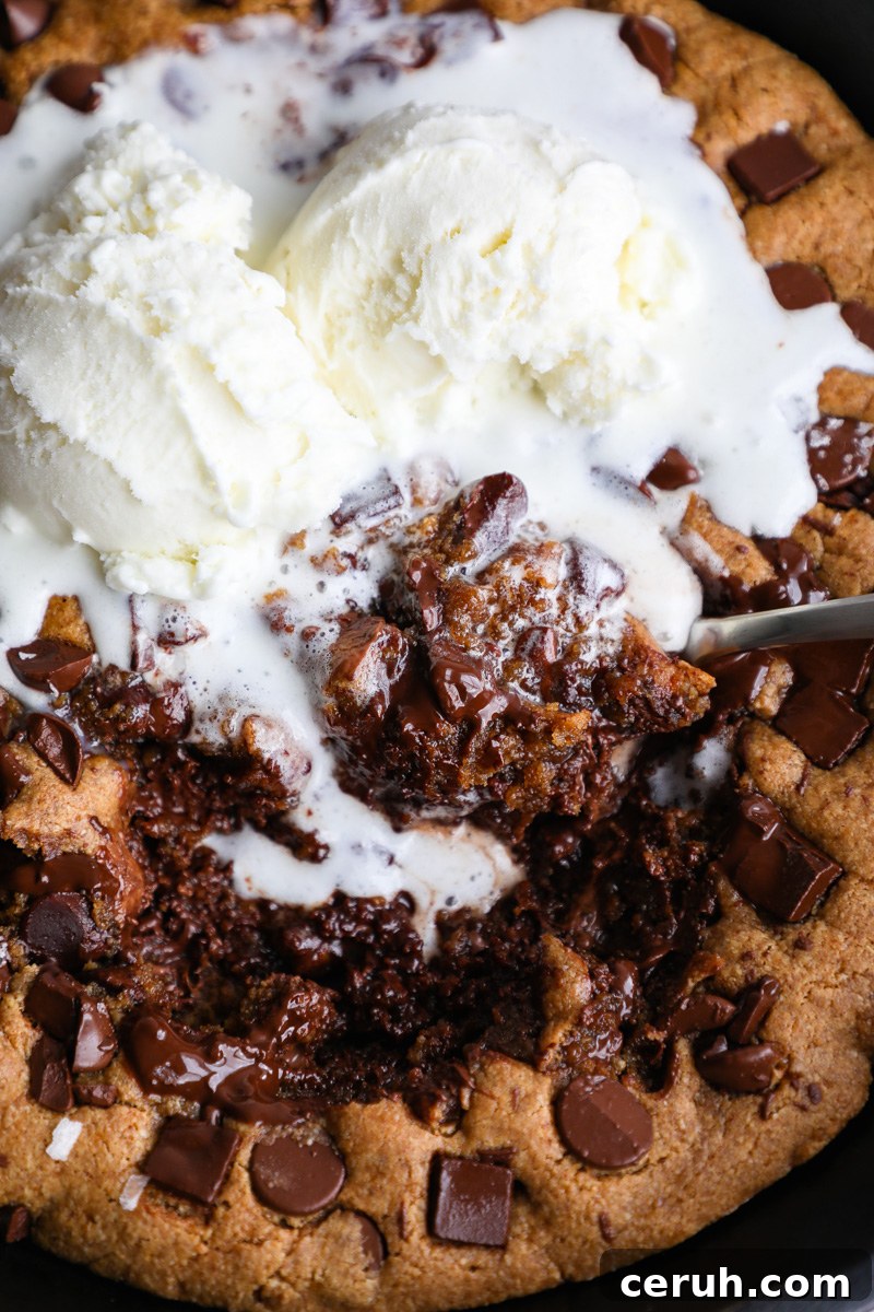 A metal spoon digging into a cast iron skillet cookie topped with vanilla ice cream that's starting to melt