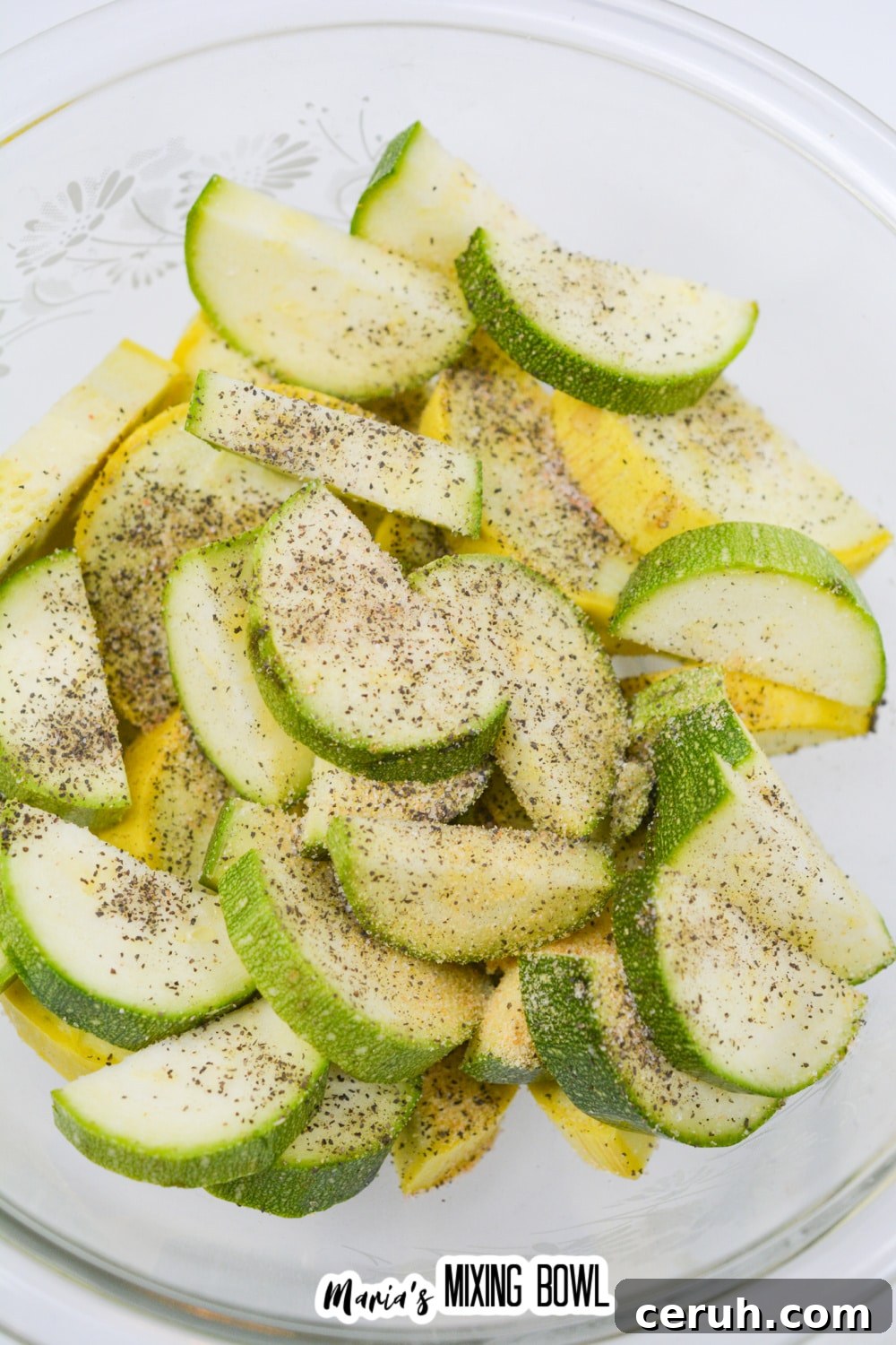 sliced raw zucchini and sliced yellow squash seasoning with salt and pepper in a glass bowl.