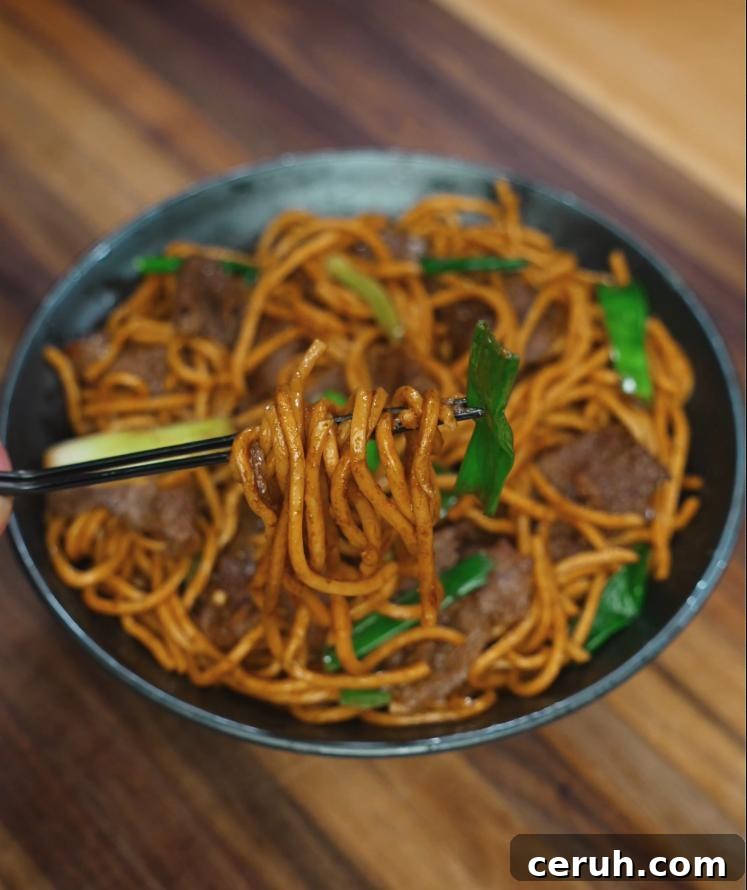 A close-up of chopsticks lifting a bite of beef lo mein, showcasing glossy noodles wrapped around stir-fried green onions and tender beef. This action shot highlights the texture and savory appeal of the dish.