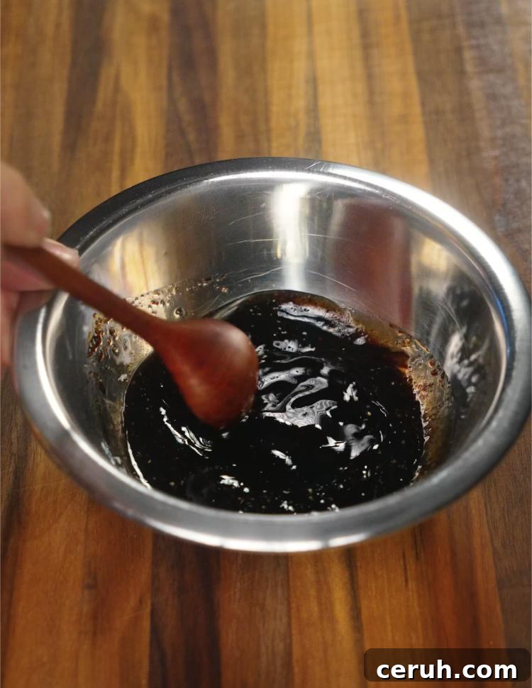 A hand mixing a glossy, dark soy sauce-based beef lo mein sauce with a wooden spoon in a metal bowl. The sauce combines soy, oyster sauce, sugar, and aromatics to deliver rich umami flavor to the noodles.