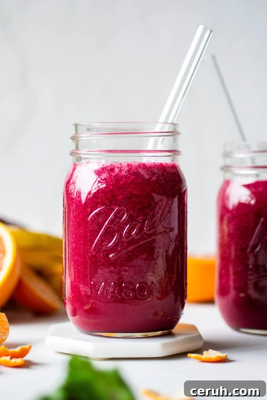 Straight on shot of 2 glass ball jars filled with a vibrant pink / red glowing beet smoothie. On a light background surrounded by fresh cut fruit. 