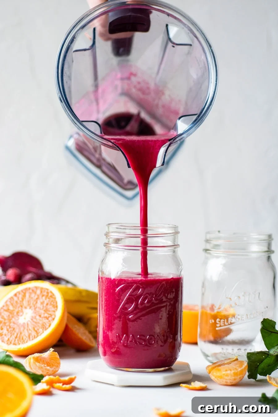 Straight on shot of a blender container pouring bright pink red beet berry smoothie into a glass ball jar. On a light background, surrounded by fresh cut fruit and beet greens. 