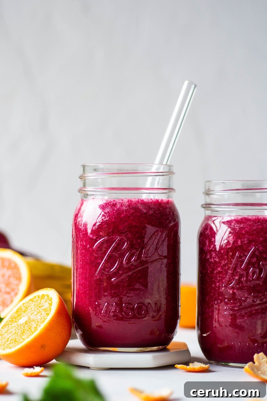 Straight on shot of 2 glass ball jars filled with a vibrant pink / red glowing beet smoothie. On a light background surrounded by fresh cut fruit and beet greens.