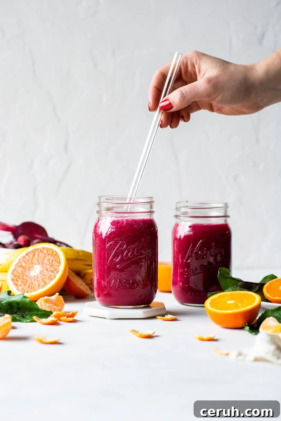 Straight on shot of 2 glass ball jars filled with a vibrant pink / red glowing beet smoothie. On a light background surrounded by fresh cut fruit, with a hand reaching into the frame to place a glass straw into one of the ball jars. 