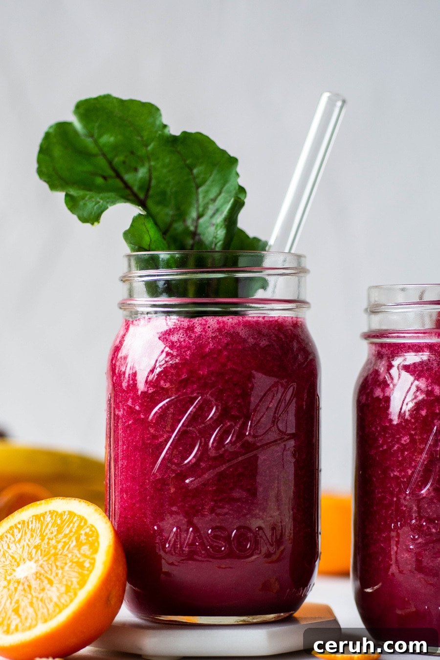 Straight on shot of 2 glass ball jars filled with a vibrant pink / red glowing beet smoothie. One jar has a beet green tucked into the jar. On a light background surrounded by fresh cut fruit. 