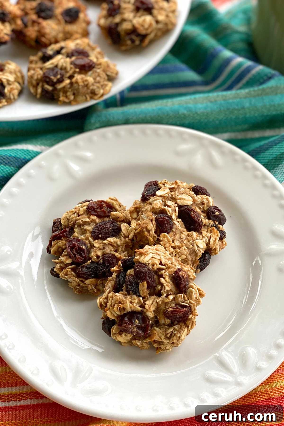 Three oatmeal banana cookies with raisin on a plate with more on a platter behind it.