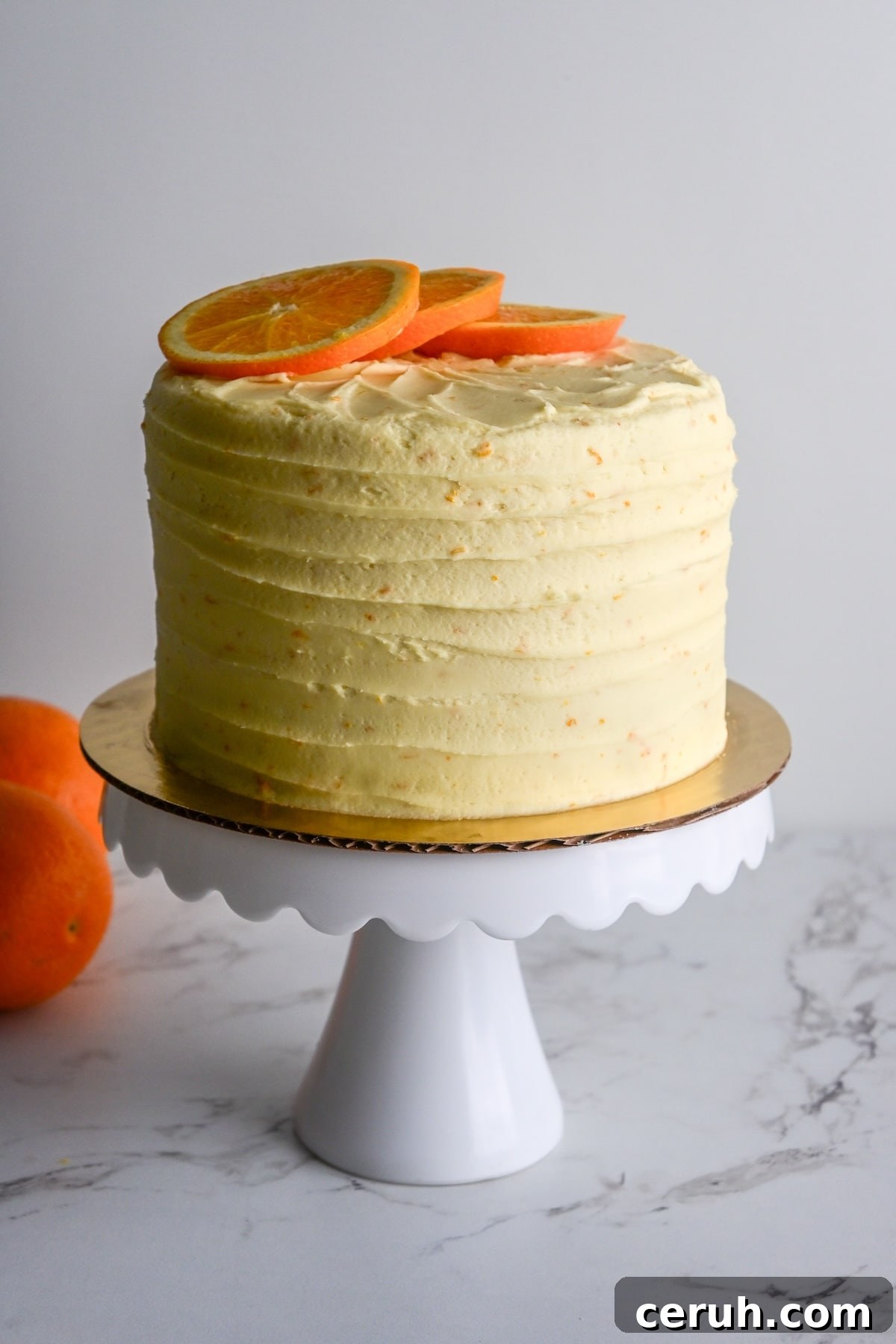 three layered orange and almond cake frosted with orange buttercream on a white pedestal with orange slices on top.