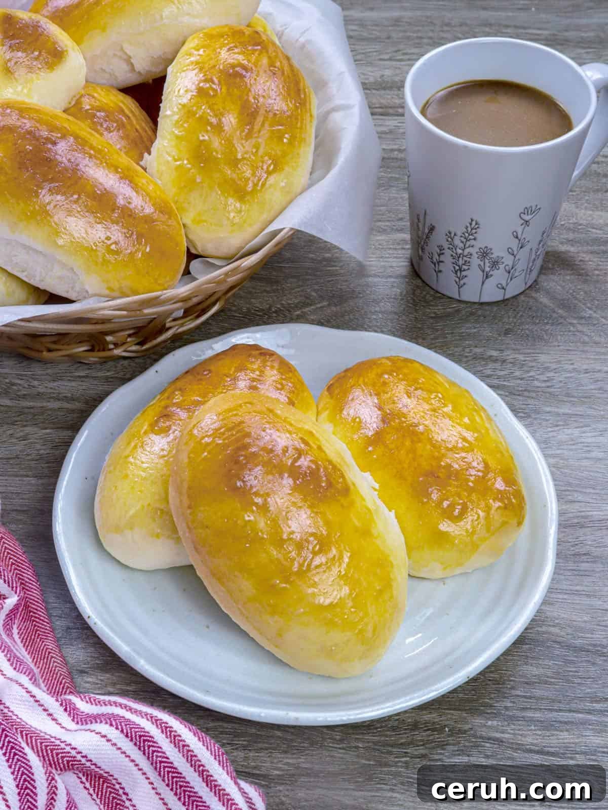 Yema Bread Rolls on a white plate with a cup of coffee and a basket of rolls in the background.