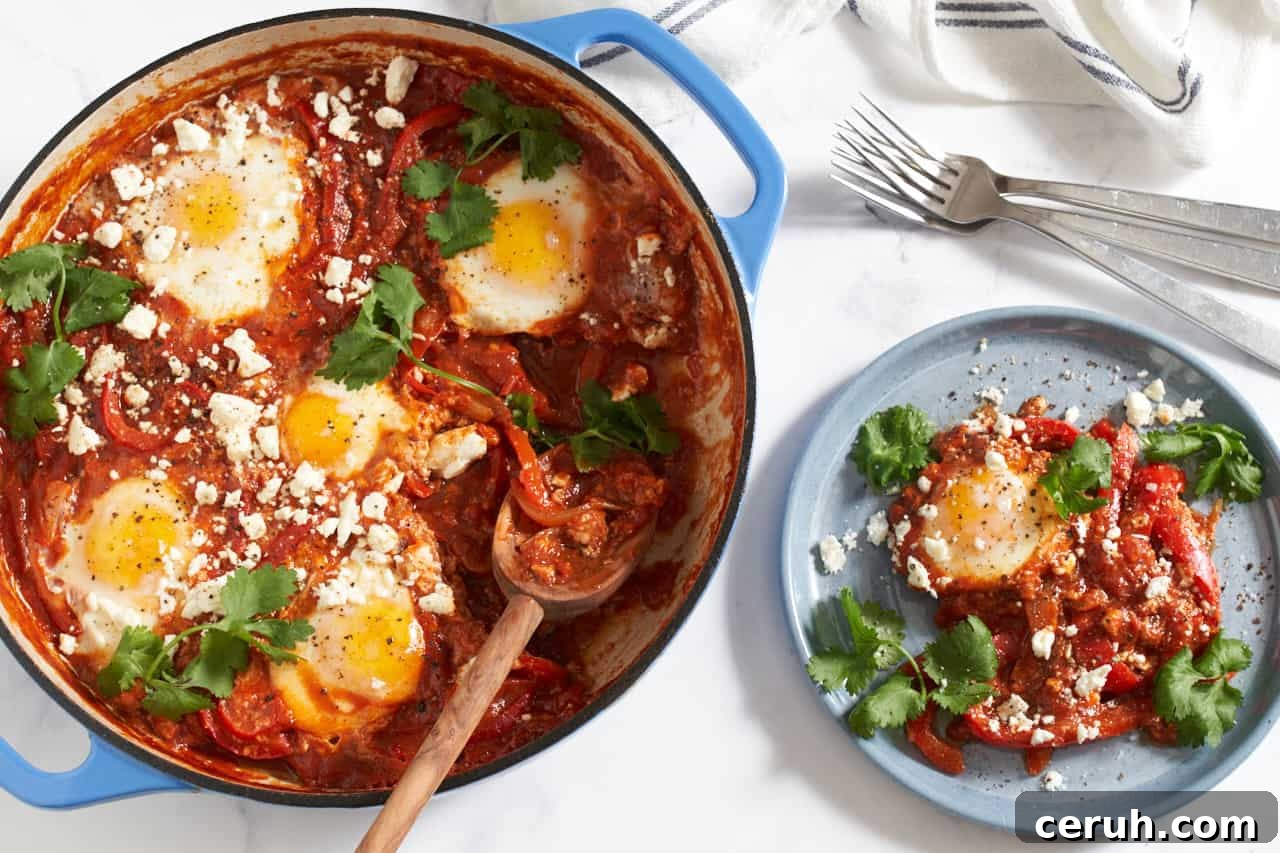 Shakshuka topped with feta and cilantro in a blue pan with a wooden spoon in it. A portion of shakshuka is on a blue plate to the right, forks and a blue and white striped towel are in the upper right corner.