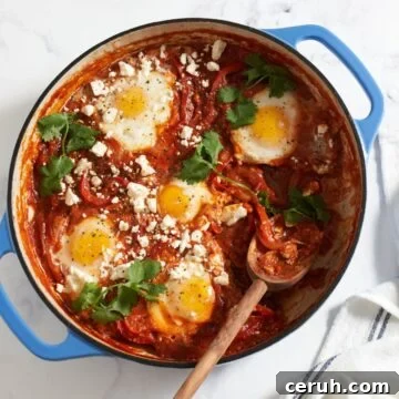 Shakshuka with feta and cilantro in a blue pan with a wooden spoon in it. A blue and white striped towel is on the right.