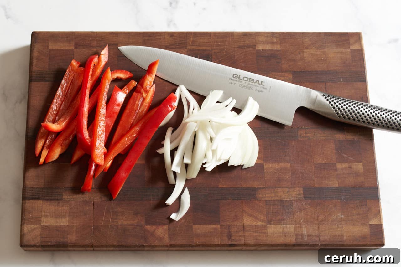 A knife on a cutting board with thinly sliced red bell peppers and onions.