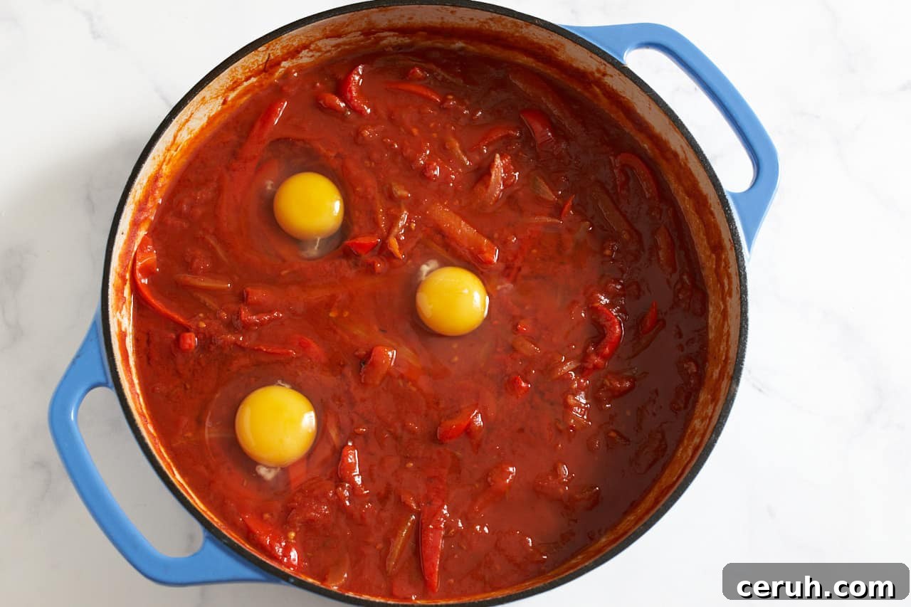 Raw eggs in shakshuka sauce in a pan with blue handles.